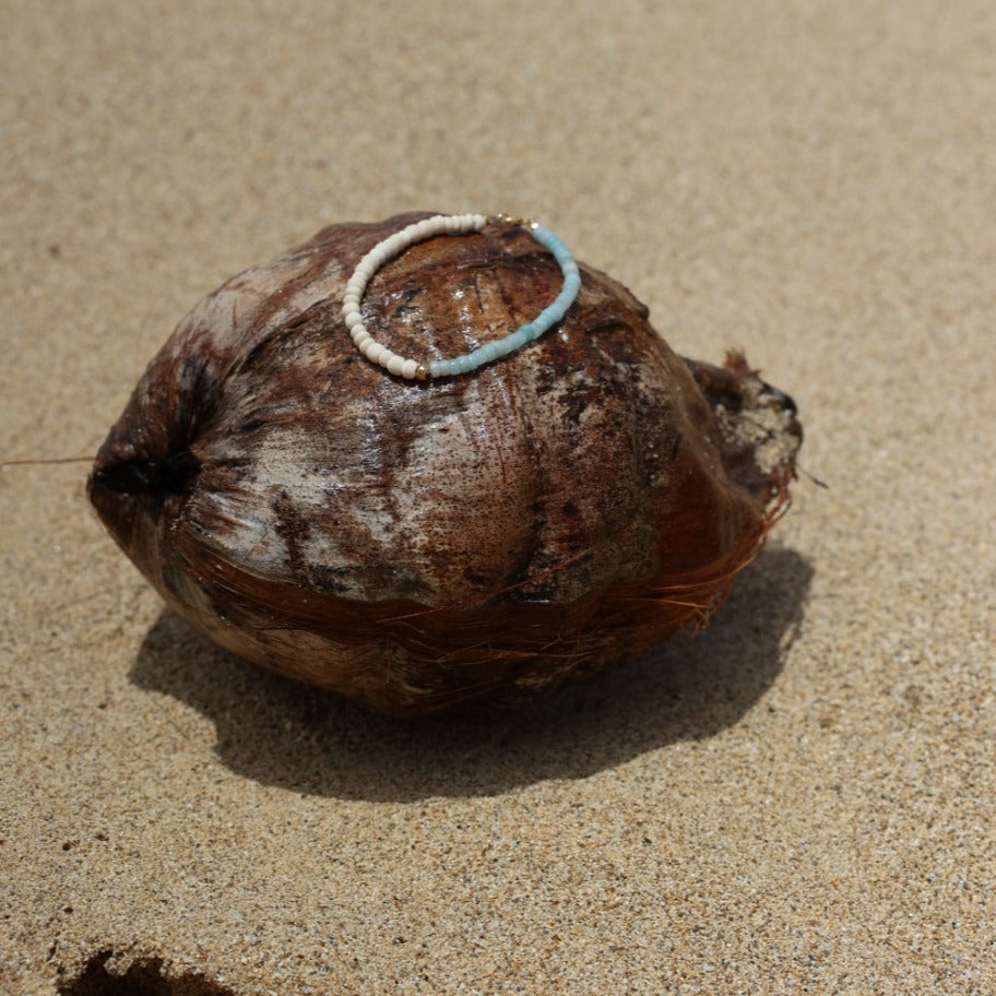 A natural brown coconut shell with a small white and light blue ring attached, lying on a sandy beige surface with a hard shadow cast to the right.