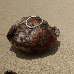 A natural brown coconut shell with a small white and light blue ring attached, lying on a sandy beige surface with a hard shadow cast to the right.