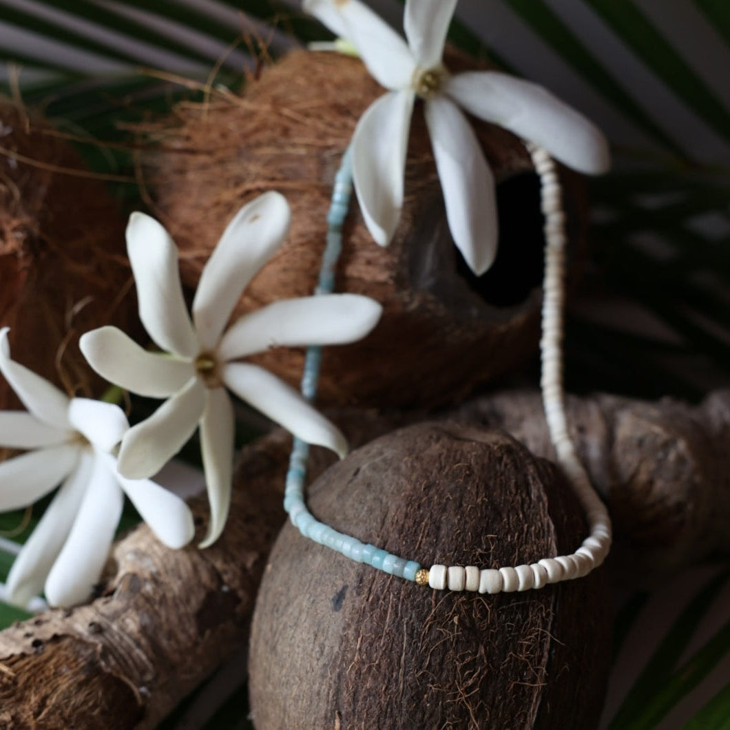 A beaded necklace with light blue and white beads draped over brown coconuts and white flowers, styled with natural wood and green leaves.