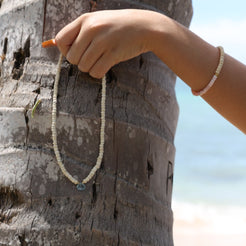 A close-up of a person's hand holding a delicate white beaded necklace against a weathered wooden barrel outdoors.