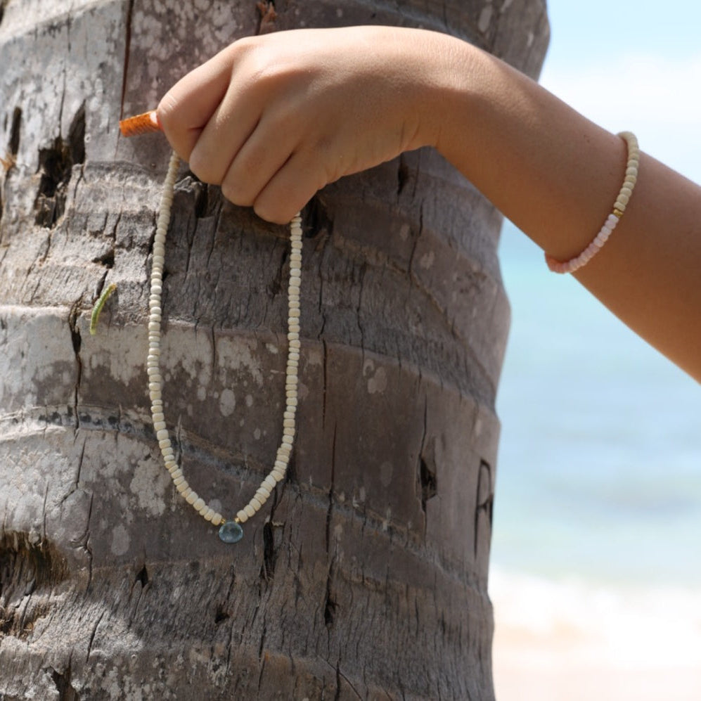 A close-up of a person's hand holding a delicate white beaded necklace against a weathered wooden barrel outdoors.