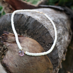A delicate beaded necklace with a white cord and a faceted purple crystal charm, styled on a textured tree trunk outdoors.