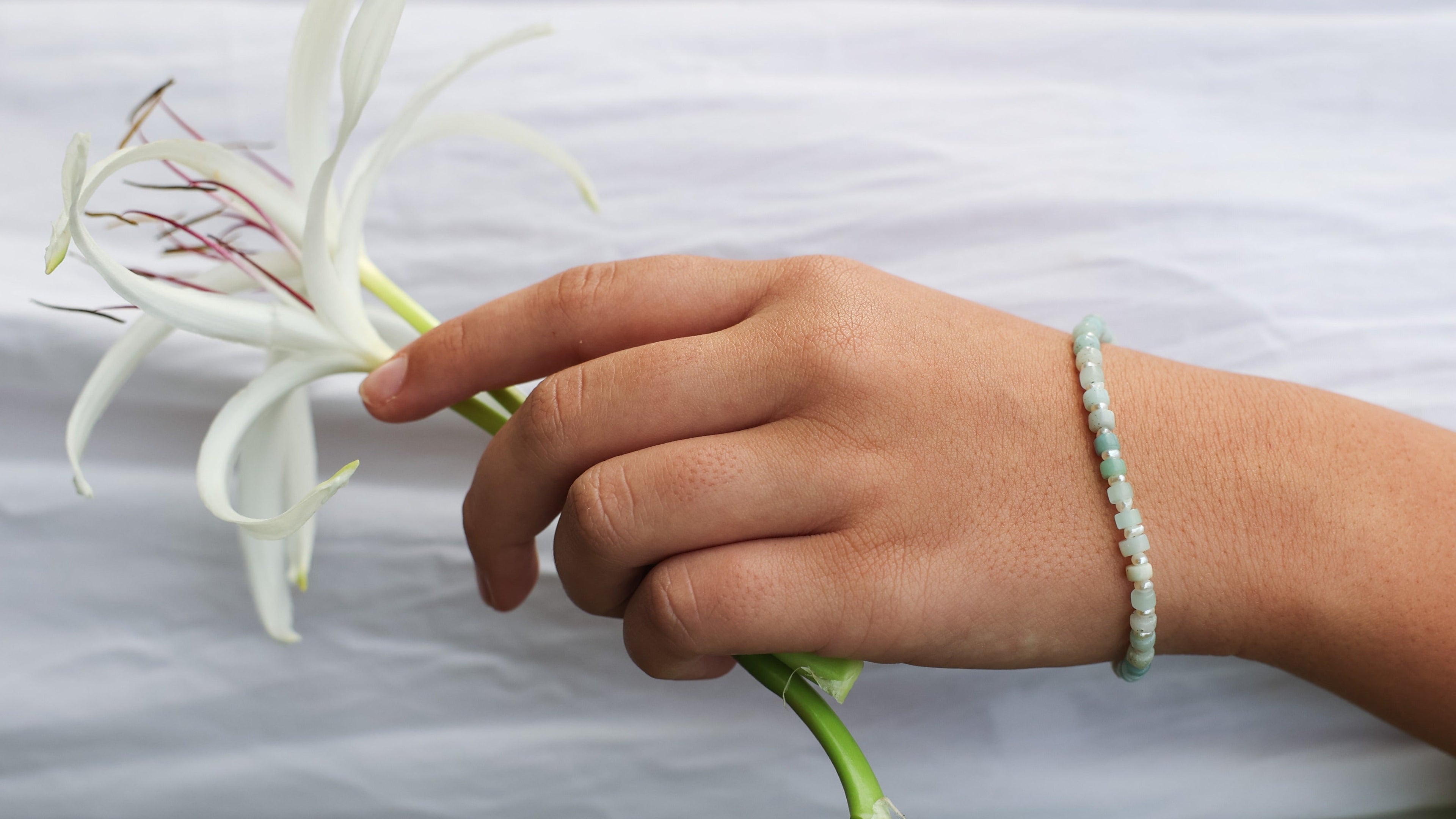 A close-up of a hand holding a white lily flower against a white fabric background, wearing a light green beaded bracelet.